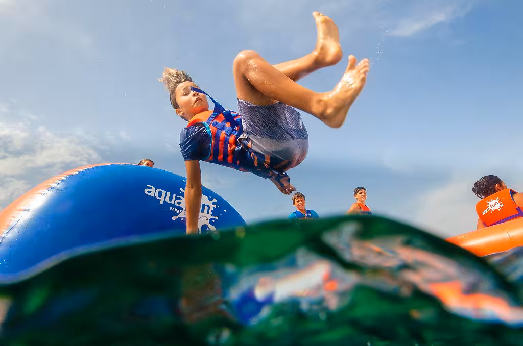 A kid in a life vest flips mid-air off an inflatable waterpark obstacle into the open water on a sunny day.