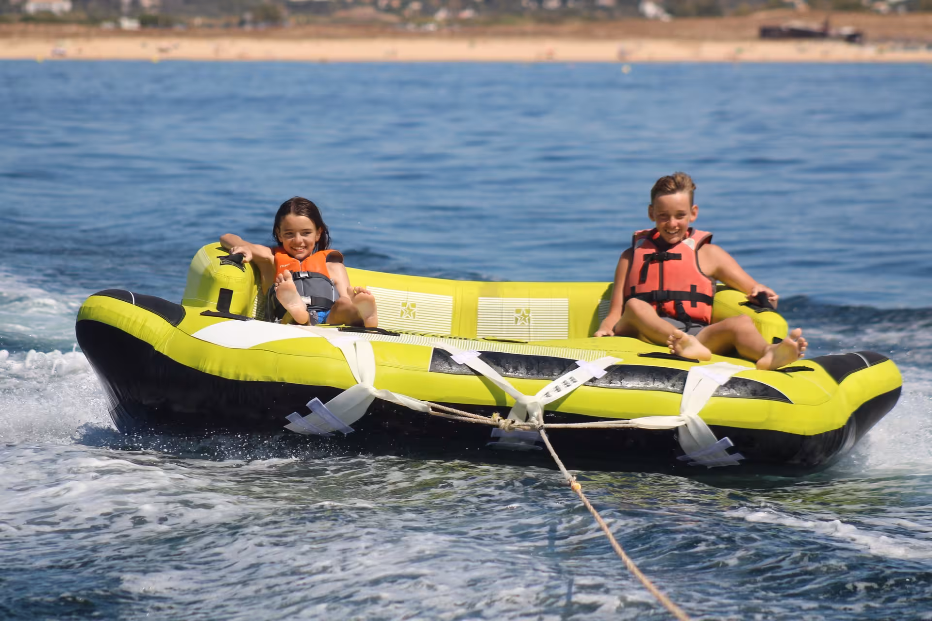 Two kids smiling and enjoying a fun Crazy Sofa inflatable tube ride on calm blue ocean waters