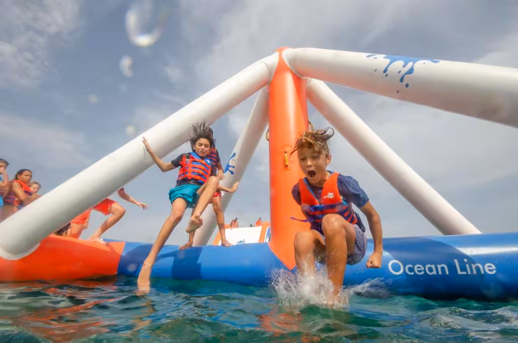 Excited kids in life vests jump and leap off inflatable waterpark Ocean Line structures into sparkling ocean water.
