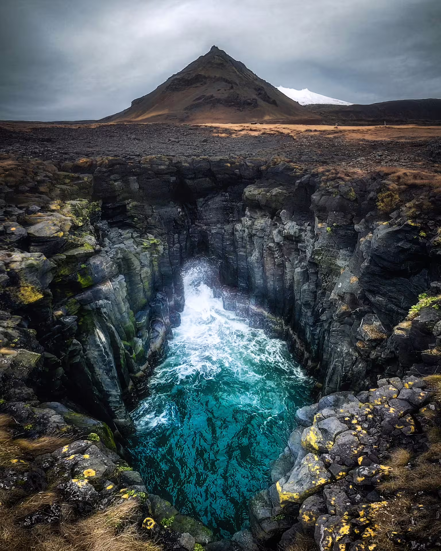 Dramatic turquoise water rushes through dark basalt rock canyon beneath iconic Kirkjufell mountain on Snaefellsnes Peninsula Iceland.