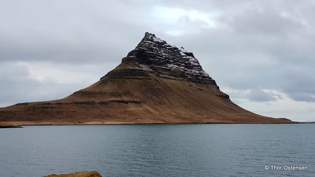 Kirkjufell mountain with snow-dusted peak reflected in calm Icelandic waters under cloudy skies