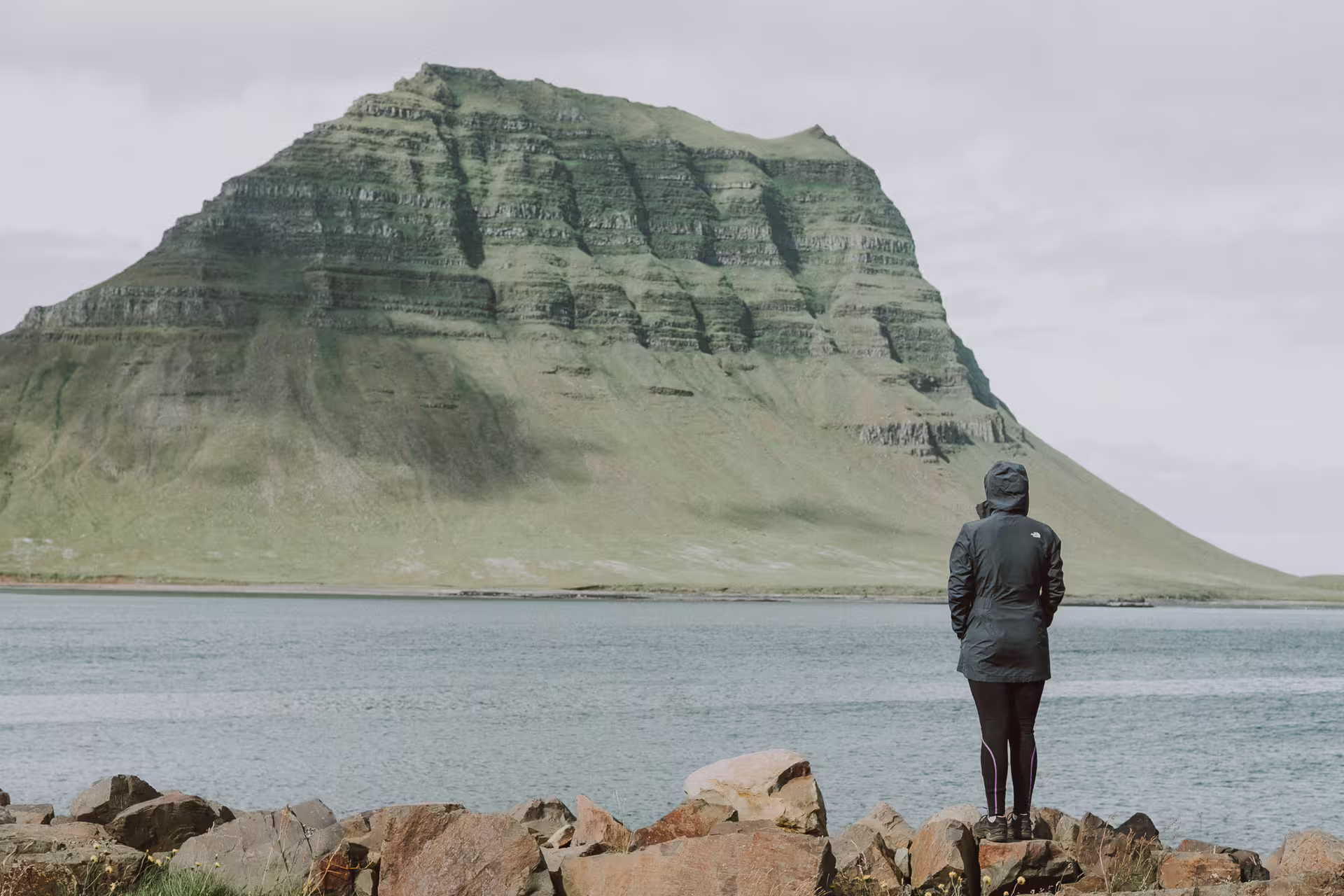 Person in North Face jacket standing on rocks gazing at Kirkjufell mountain across calm waters on Snaefellsnes Peninsula