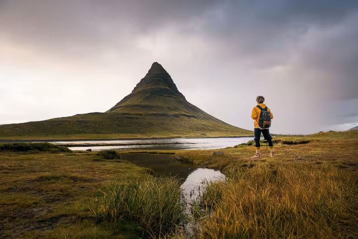 Hiker in yellow jacket with backpack gazing at Kirkjufell mountain and reflective lake on Snaefellsnes Peninsula