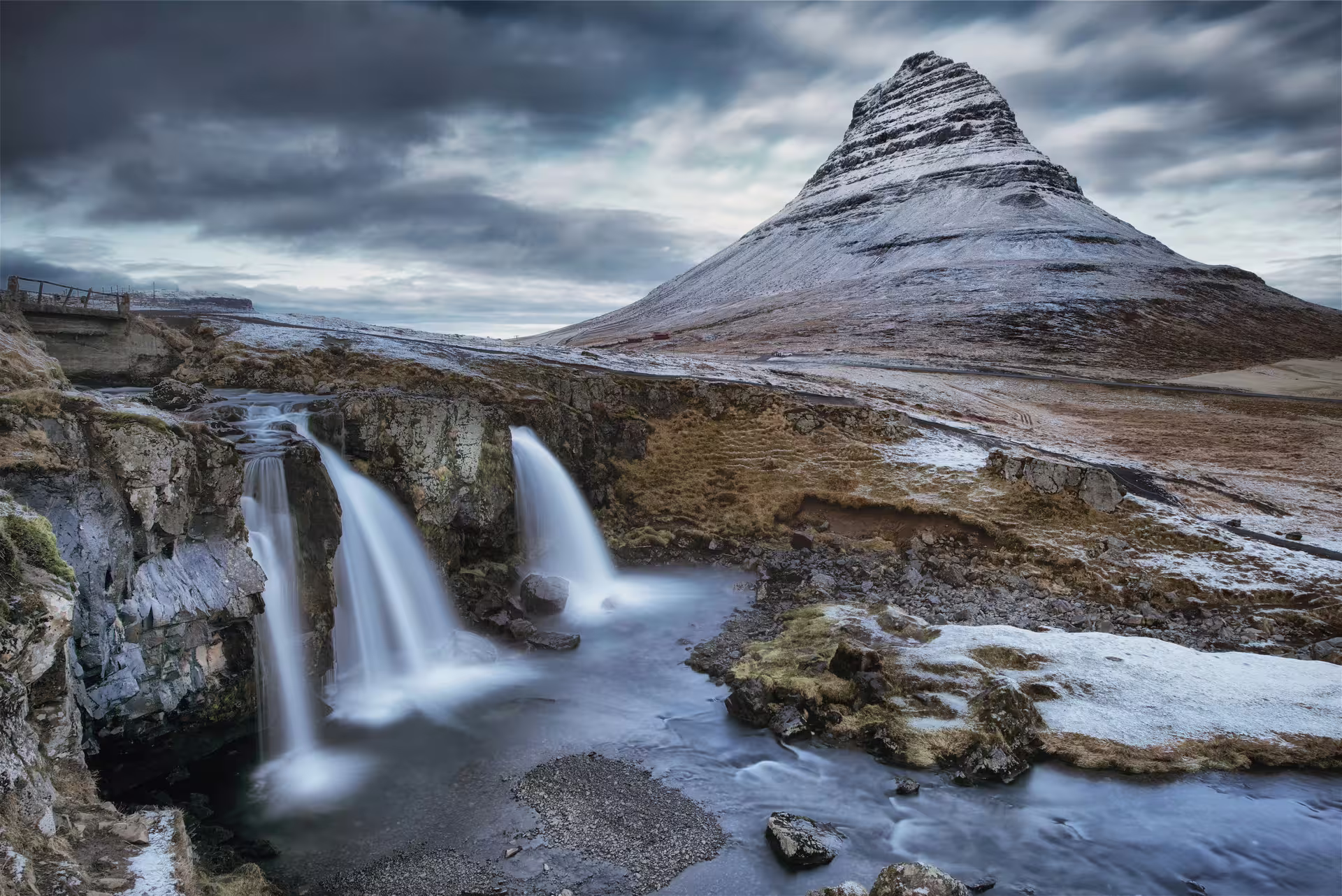 Kirkjufellsfoss waterfall cascading before snow-dusted Kirkjufell mountain under dramatic cloudy skies, Snæfellsnes Peninsula, Iceland