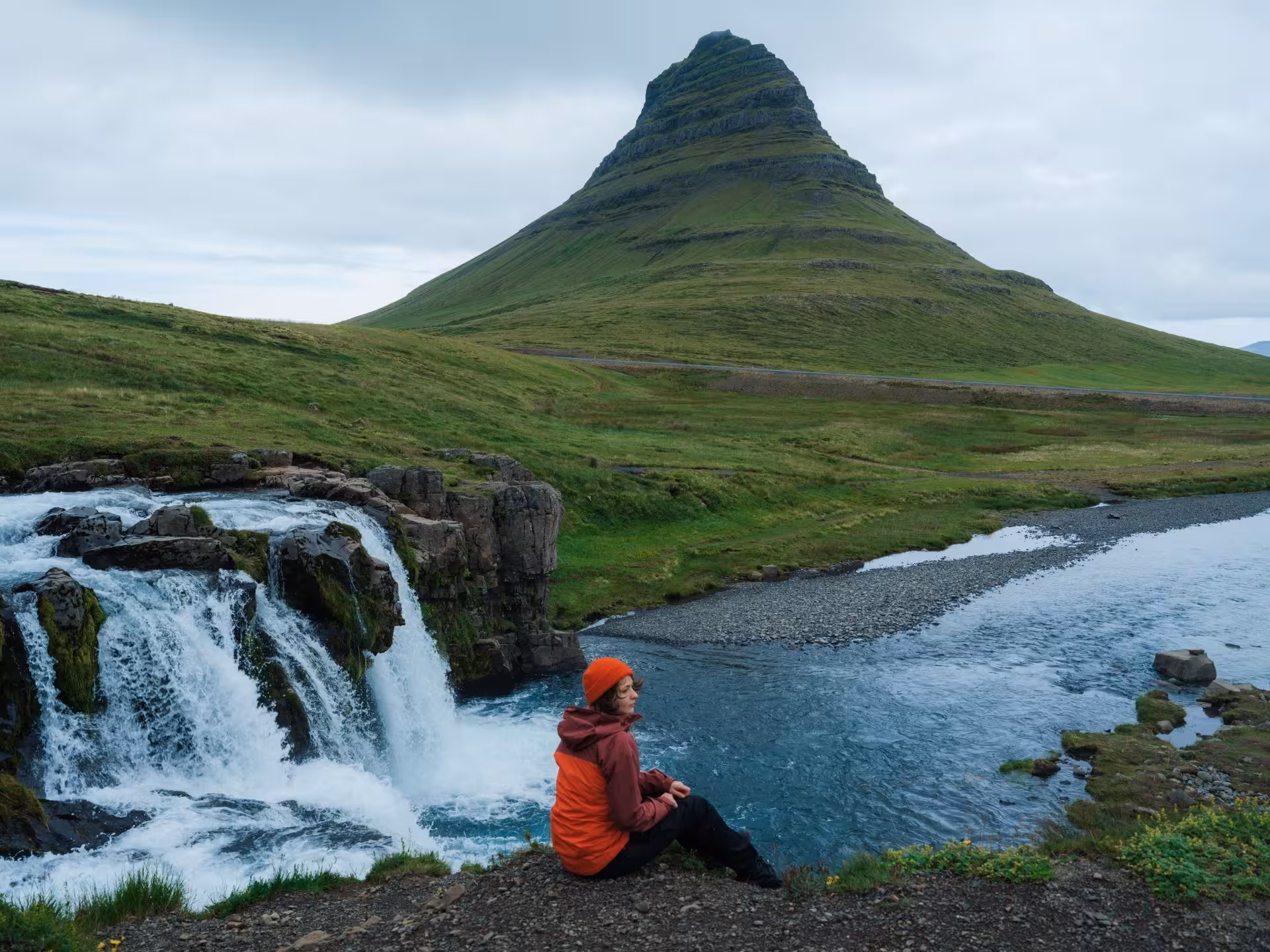 Traveler sitting beside Kirkjufellsfoss waterfall with iconic Kirkjufell mountain in background, Snaefellsnes Peninsula, Iceland