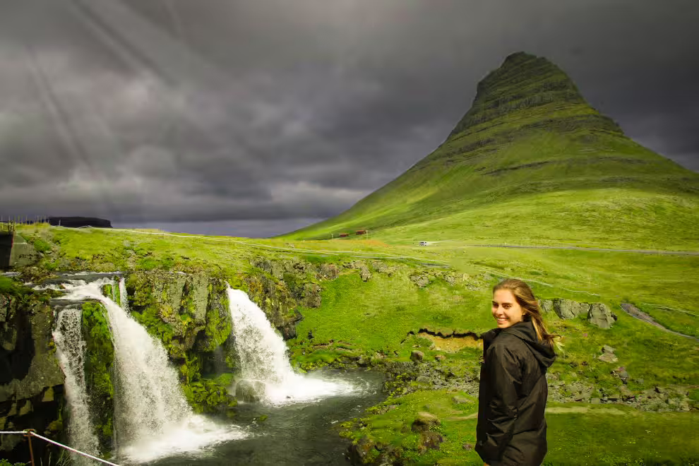 Woman smiling by Kirkjufellsfoss waterfall with iconic Kirkjufell mountain and dramatic skies, Iceland
