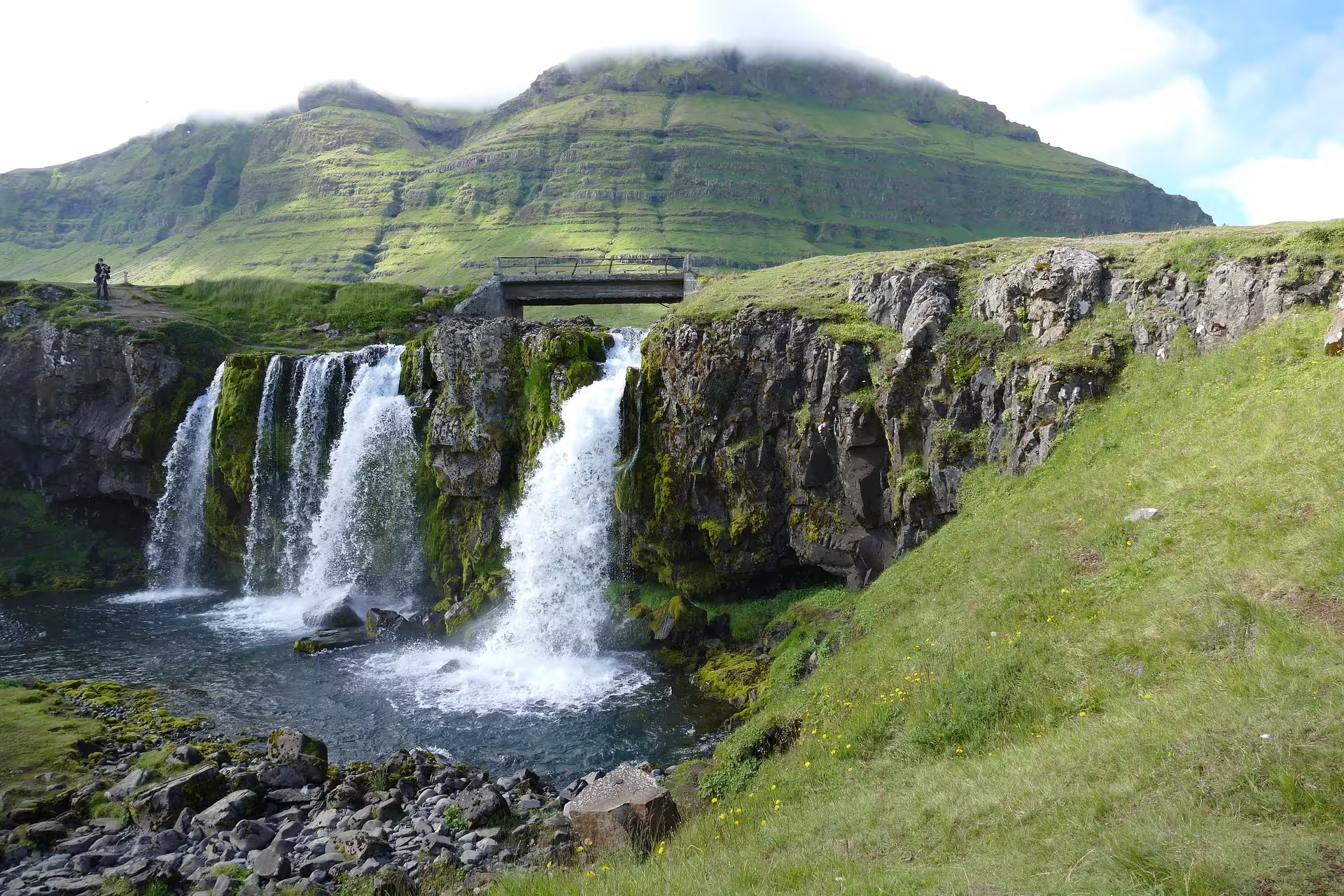 Kirkjufellsfoss waterfall cascading over mossy cliffs with green mountain and bridge on Snaefellsnes Peninsula