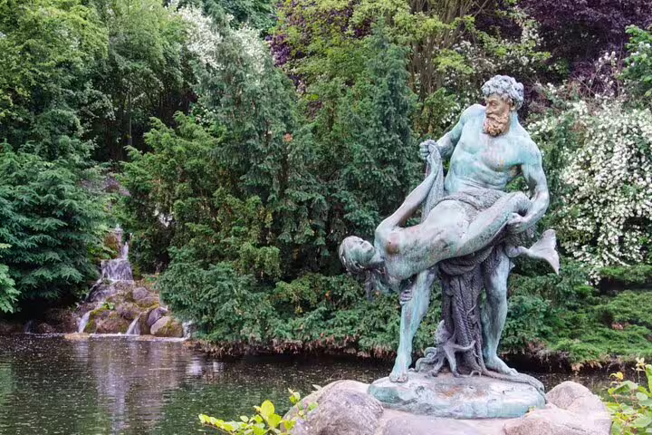 Bronze statue of a bearded man carrying a figure beside a garden pond with waterfall in Berlin park