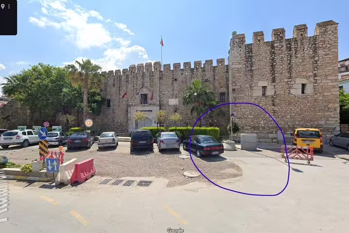 Historic stone castle near Kusadasi Port surrounded by parked cars and lush greenery under a bright blue sky.