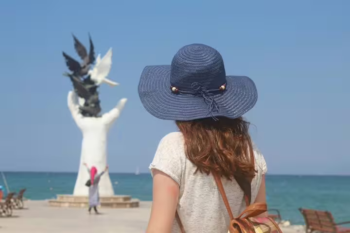 Woman in blue sun hat facing a white hand sculpture releasing birds on Kusadasi seafront