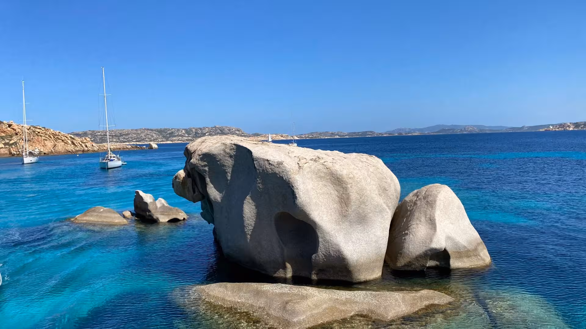 Sailboats anchored near massive rock formations in crystal-clear waters of the La Maddalena Archipelago, ideal for tours.