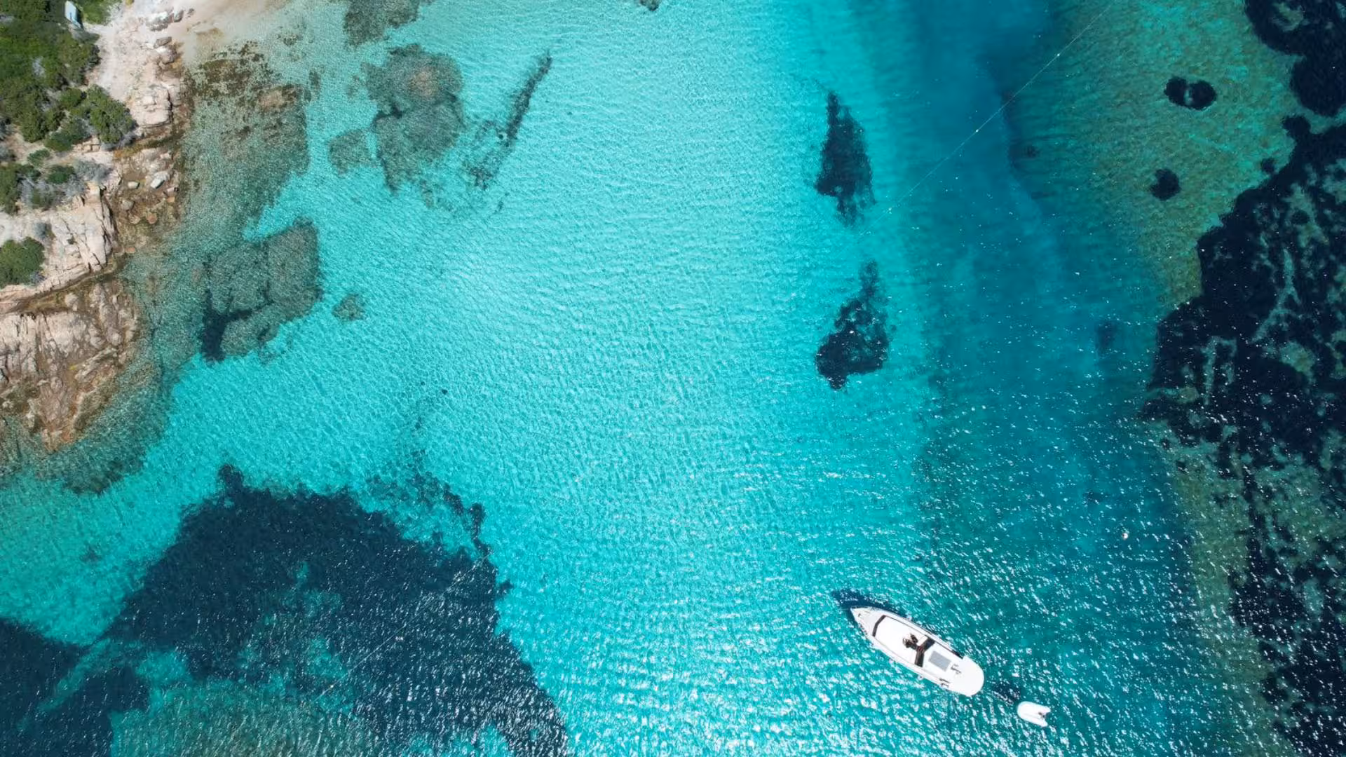 Aerial view of a white boat cruising through the crystal-clear waters of La Maddalena Archipelago's rocky coastline.