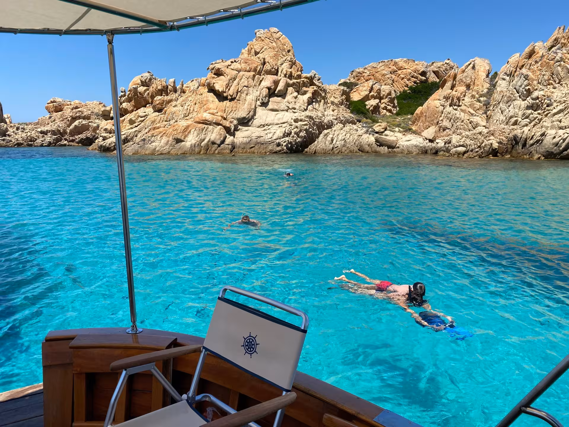 Snorkelers explore turquoise waters by rugged rocks on a wooden boat tour in La Maddalena archipelago.