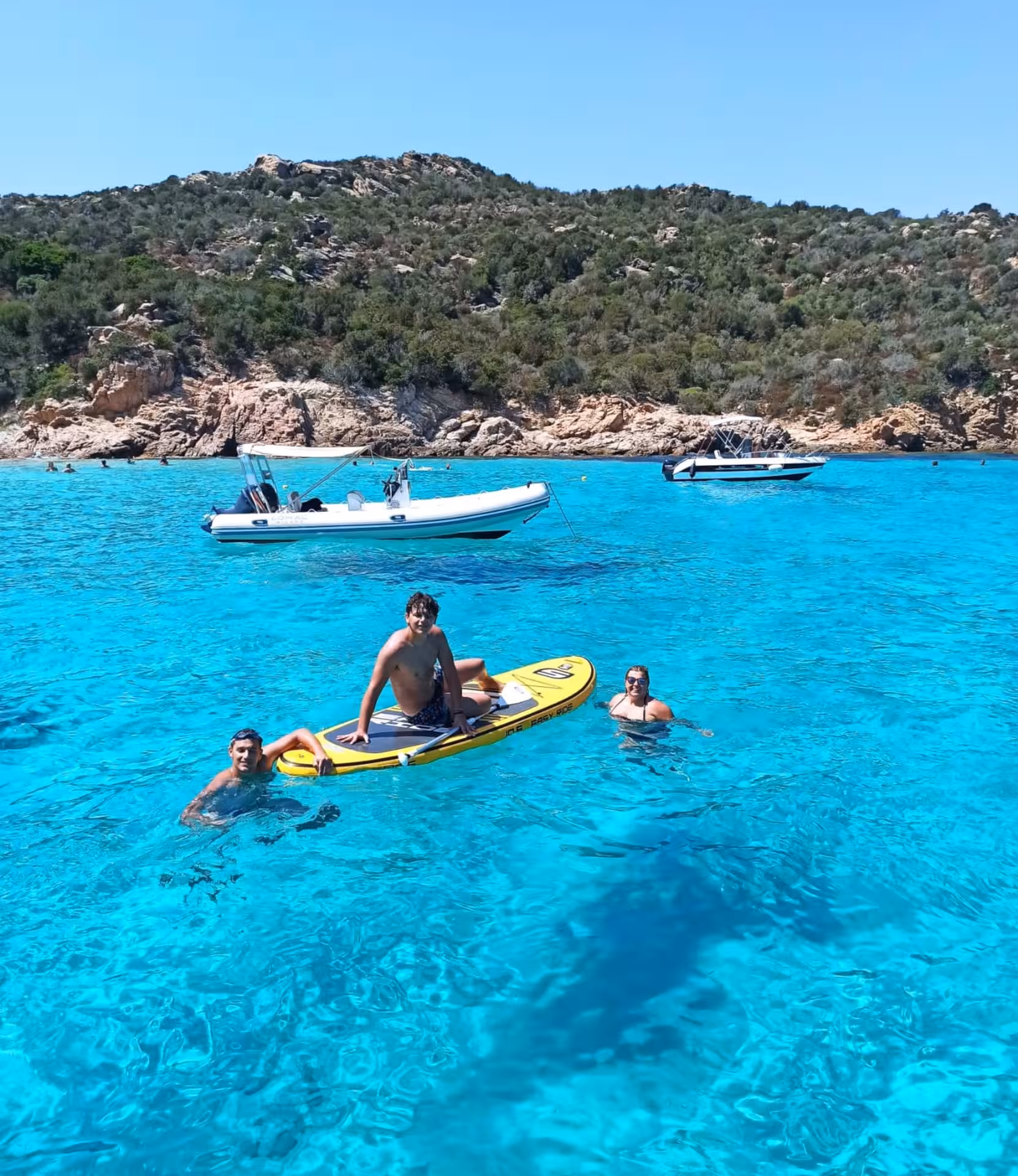 Group enjoying paddleboarding and swimming in the vibrant blue waters of La Maddalena Archipelago, with boats nearby.