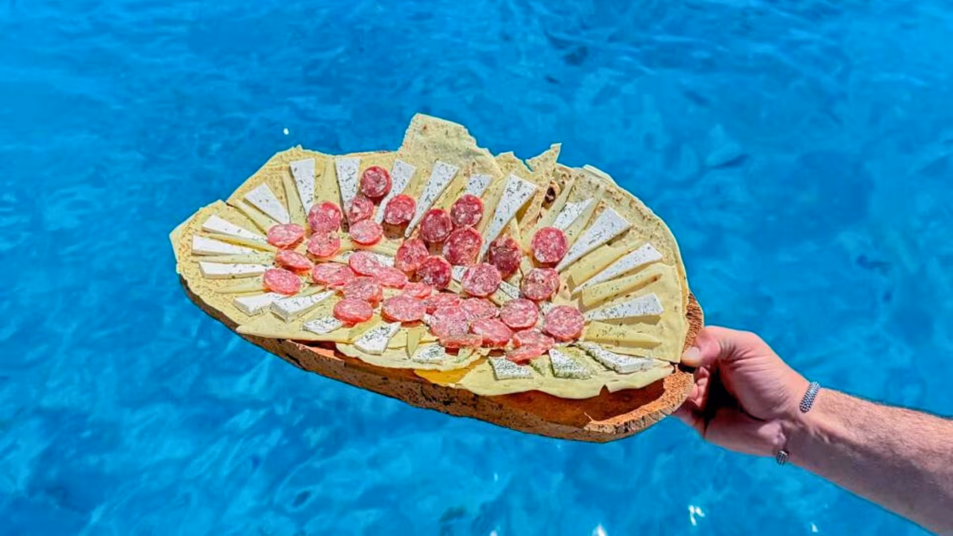 Delicious platter with meats and cheeses held over turquoise waters during La Maddalena Archipelago sailing tour.