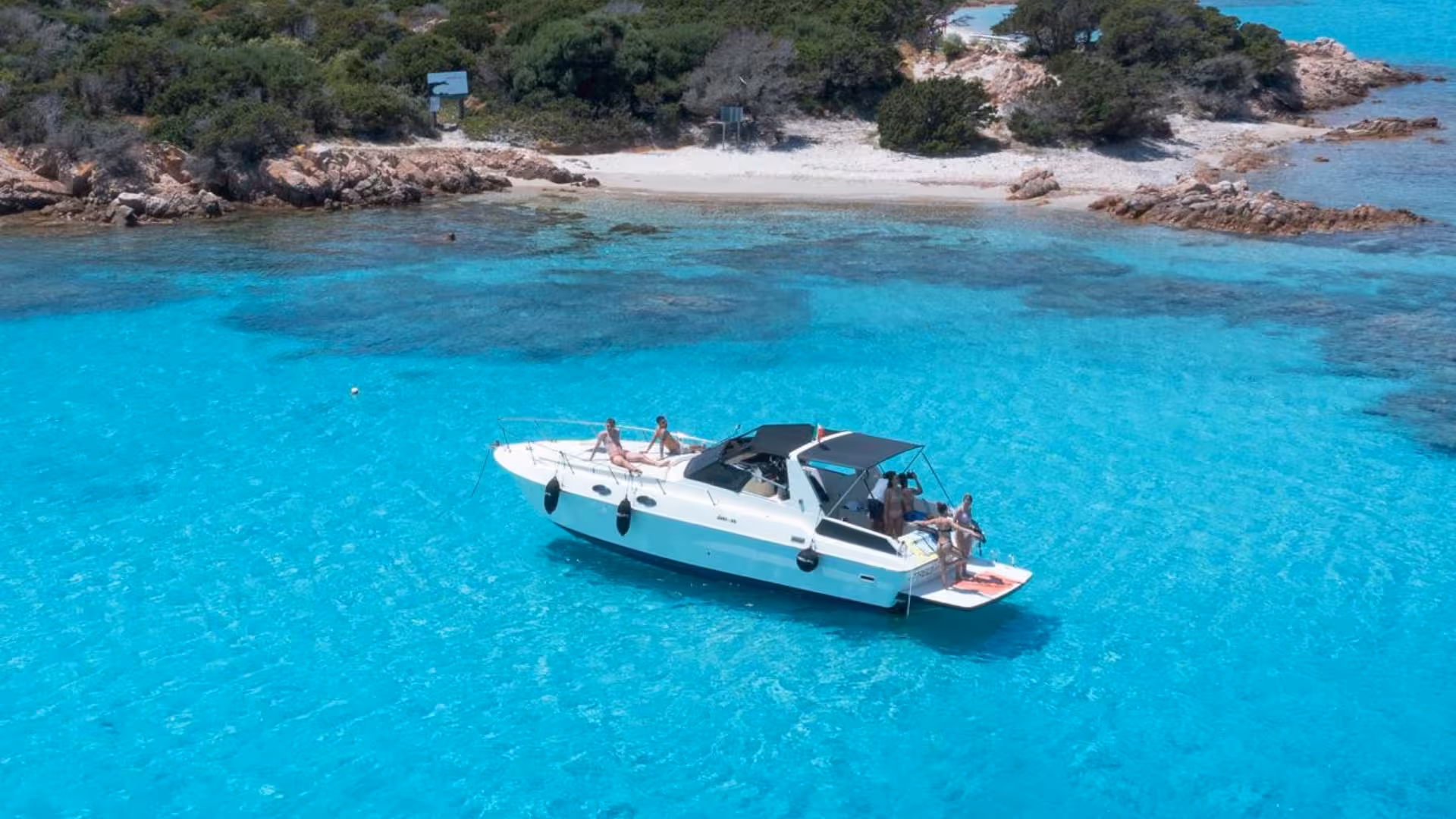 Tourists relaxing on a yacht in the crystal-clear waters of La Maddalena Archipelago near a sandy cove.