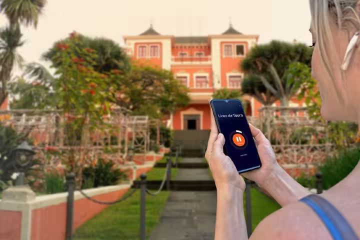 Woman holding smartphone with Liceo de Taoro audio guide app in front of red historic building in La Orotava