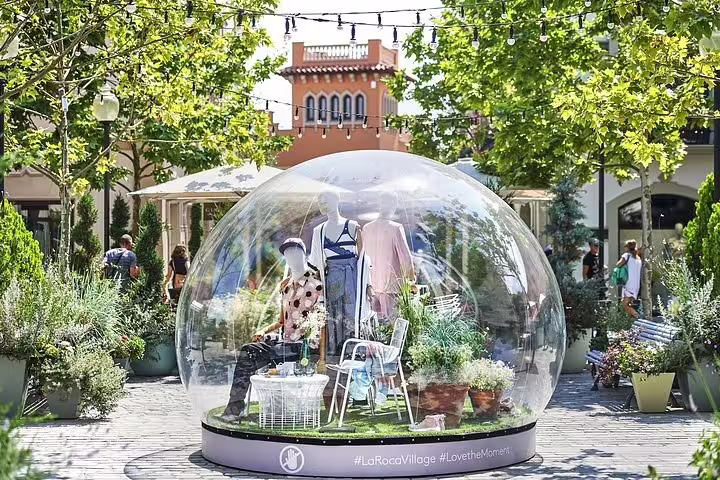 Mannequins in stylish outfits inside a transparent dome at La Roca Village, surrounded by lush greenery and shoppers.