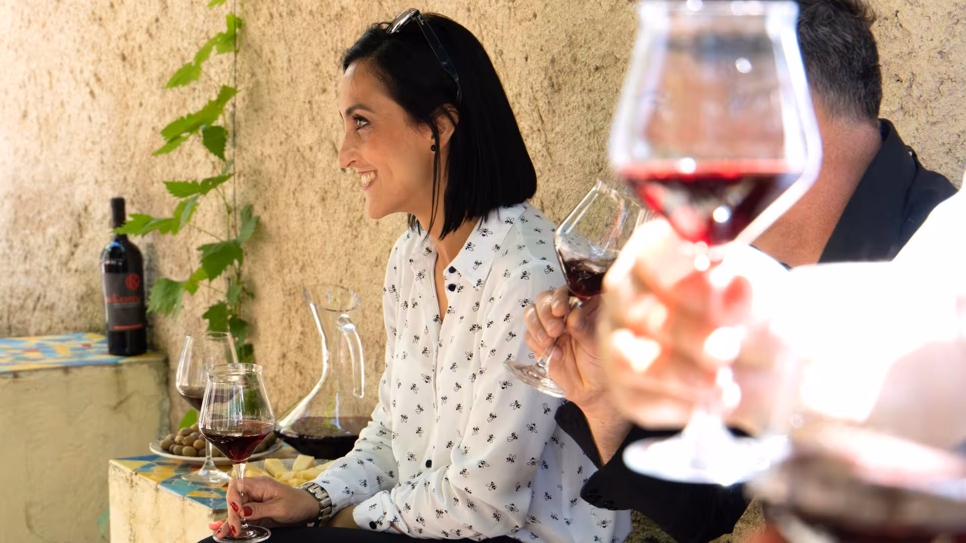 People enjoying red wine tasting at a Laconi winery, smiling and surrounded by lush greenery and wine bottles.