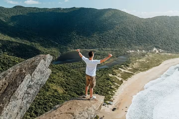 Adventurer standing on a cliff overlooking Lagoinha do Leste beach and lush forests during a hiking tour in Brazil.