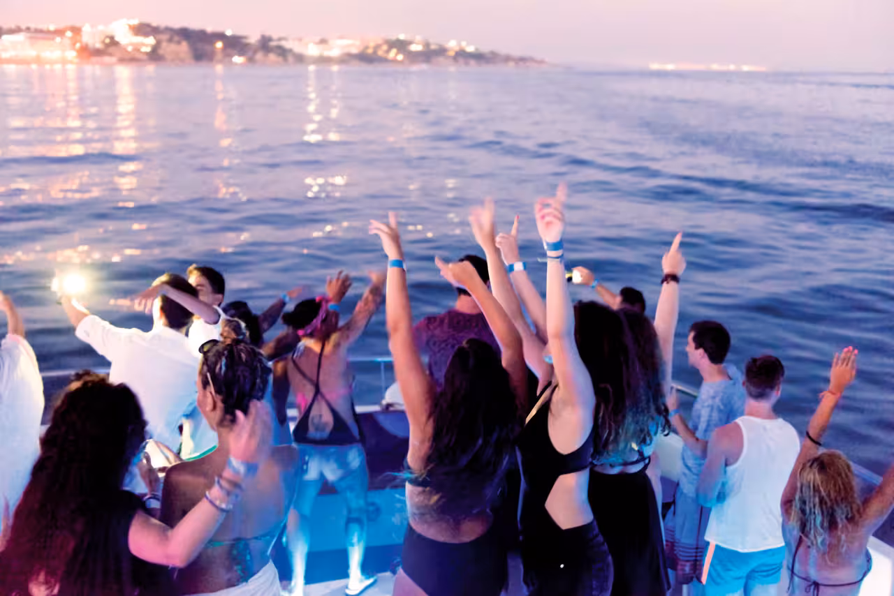 Energetic crowd dancing with hands raised on a Lagos boat party at dusk with glowing coastal lights.