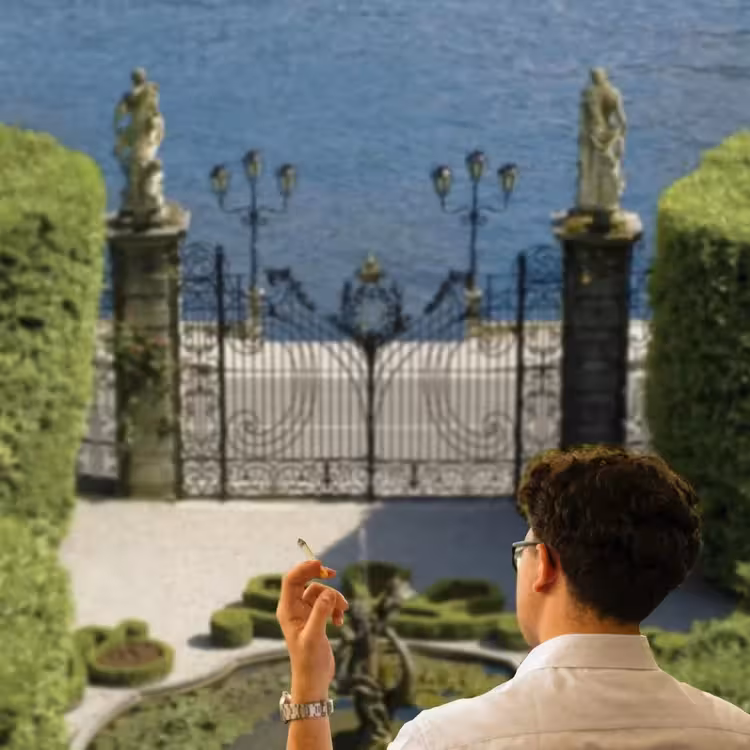 Person holding cigarette overlooking ornate iron gates and Lake Como garden landscape