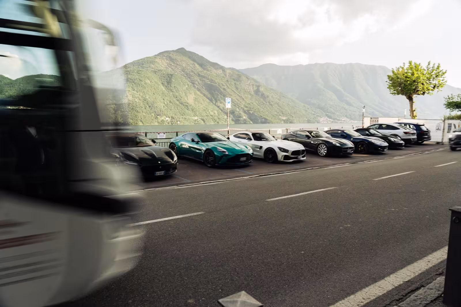 Luxury supercars including Aston Martin parked along Lake Como waterfront with alpine mountains backdrop