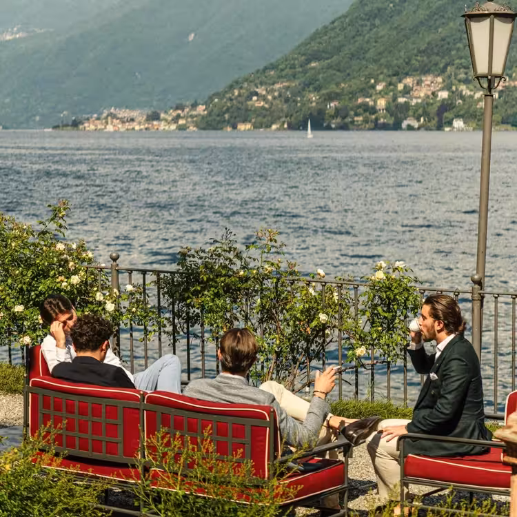 Elegantly dressed men relaxing on red outdoor sofas on a lakeside terrace at Lake Como with mountain views