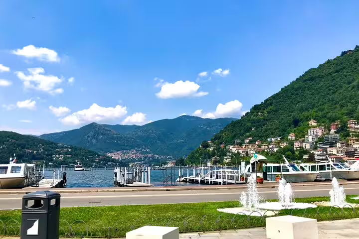 Lake Como waterfront promenade with marina, fountains, and green hillside town under blue sky