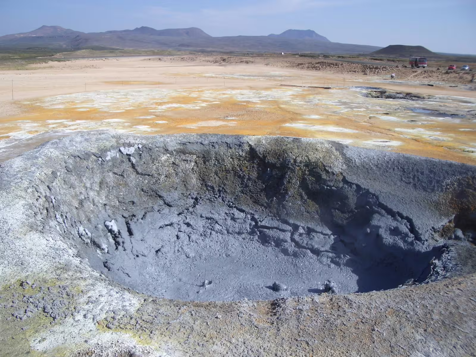 Bubbling grey mud pool at Lake Myvatn geothermal area, Iceland, with colorful sulfur deposits and volcanic mountains in background