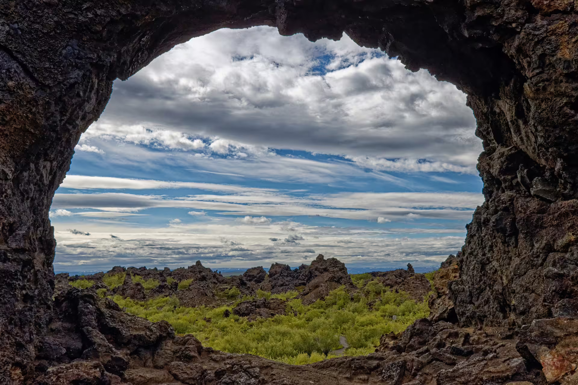View through volcanic lava rock arch at Lake Myvatn lava fields with green moss and cloudy blue sky, Iceland