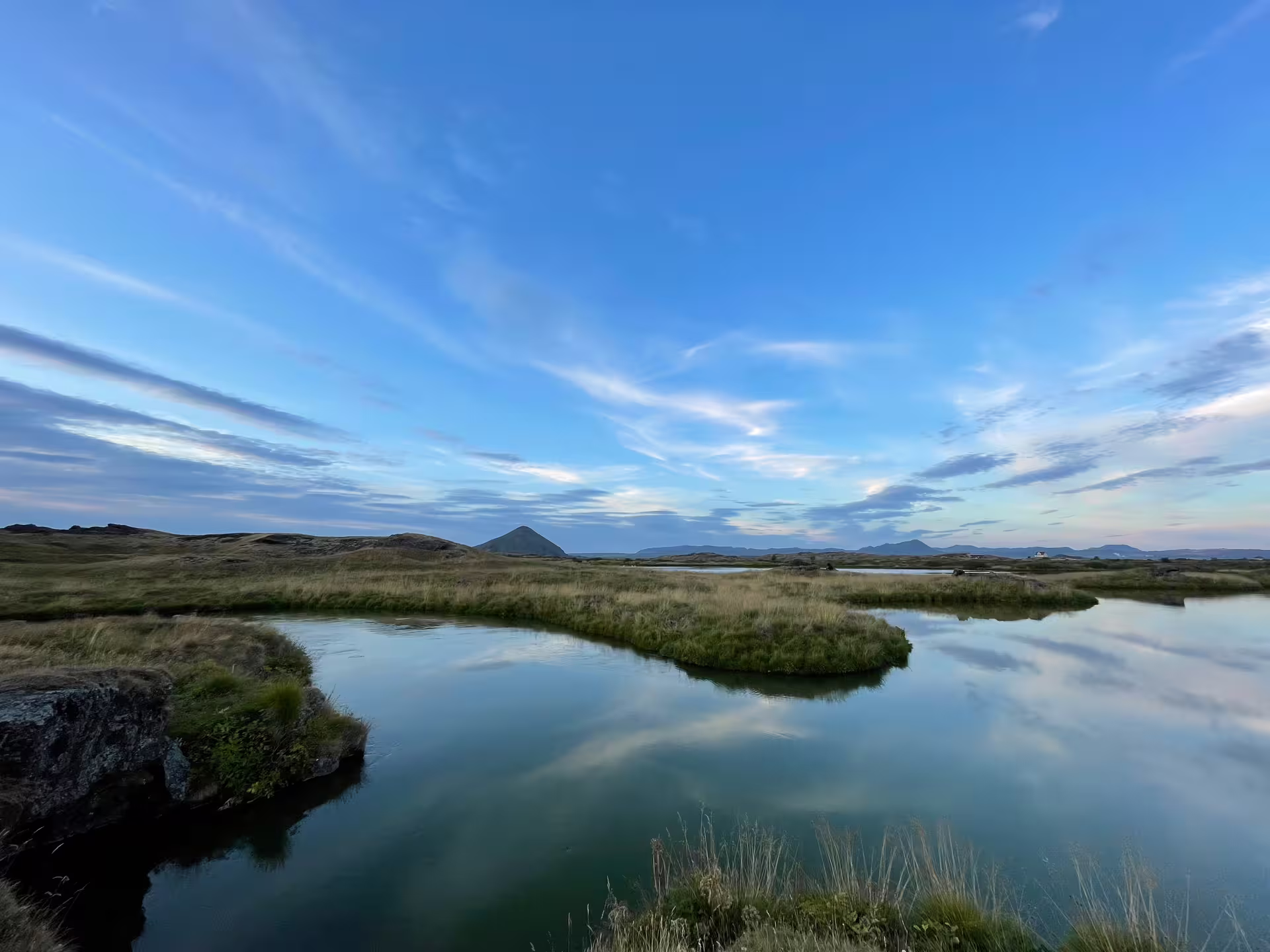 Calm reflective waters of Lake Myvatn with volcanic cone and golden grasslands under blue sky, Iceland