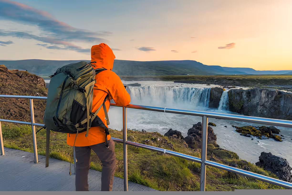 Hiker in orange jacket with backpack overlooking Godafoss waterfall at sunset, Lake Myvatn Iceland