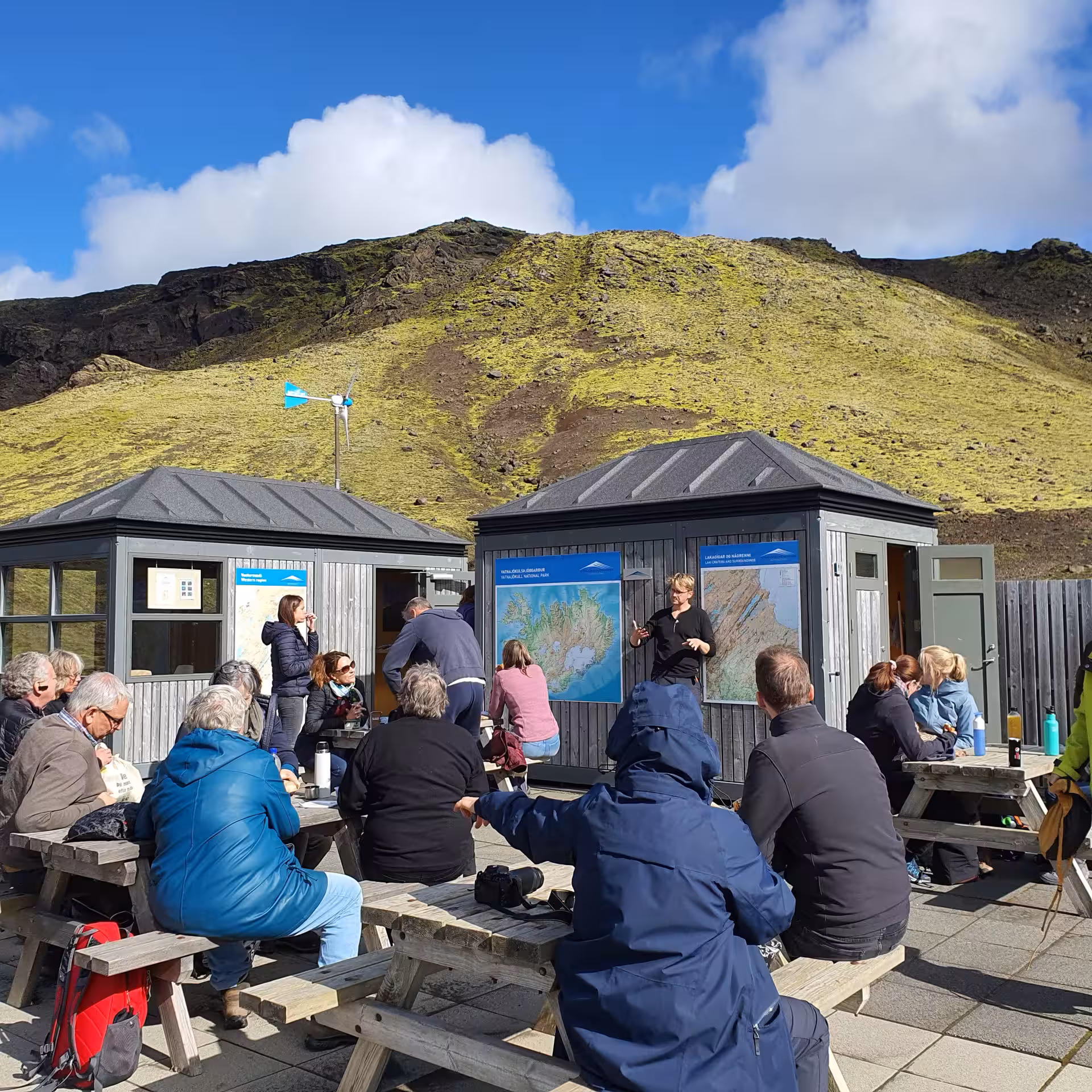 Tour guide presenting Iceland map to group of tourists at Laki Craters visitor huts with volcanic hill backdrop
