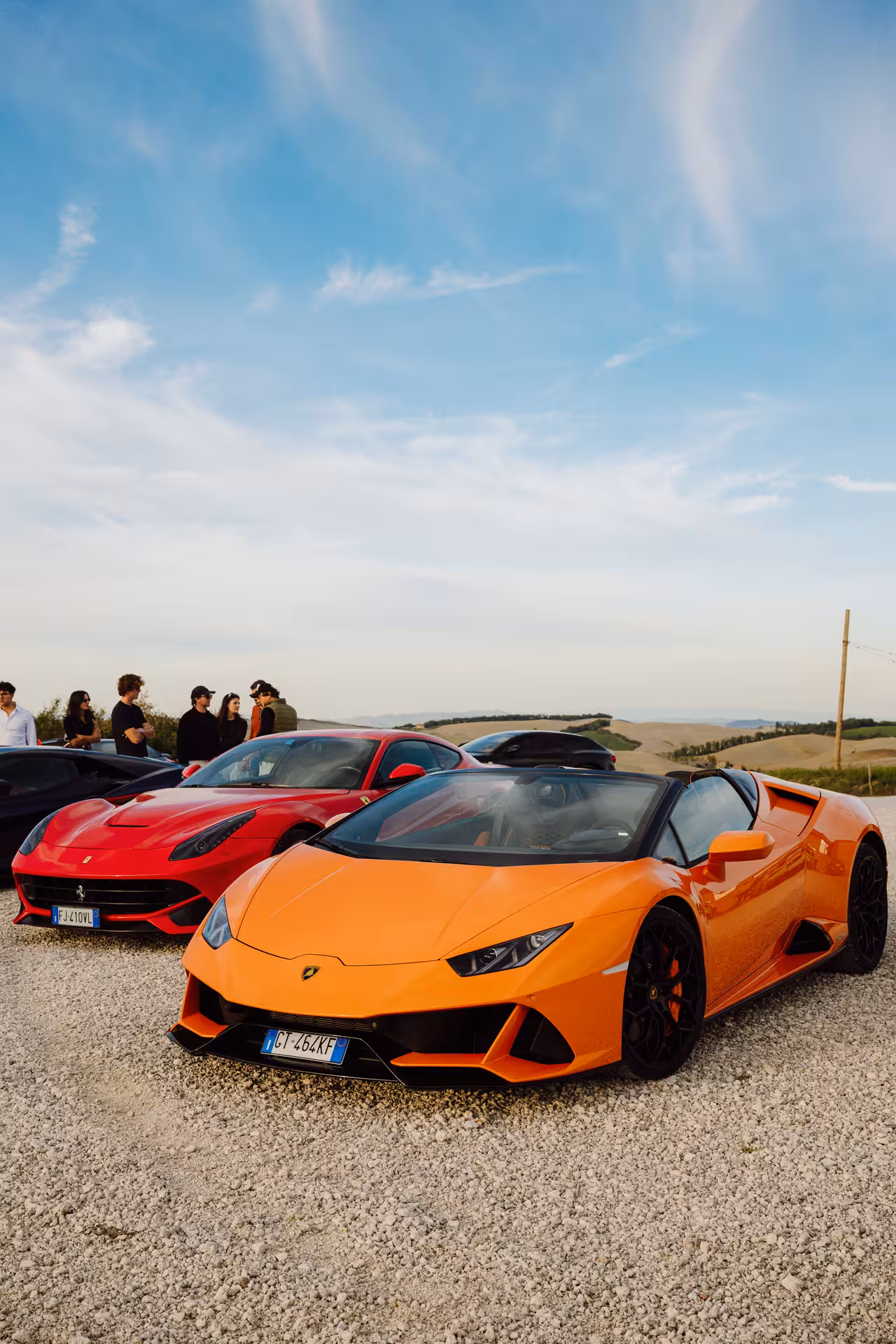 Orange Lamborghini Huracan and red Ferrari parked on gravel with Tuscan hills in background