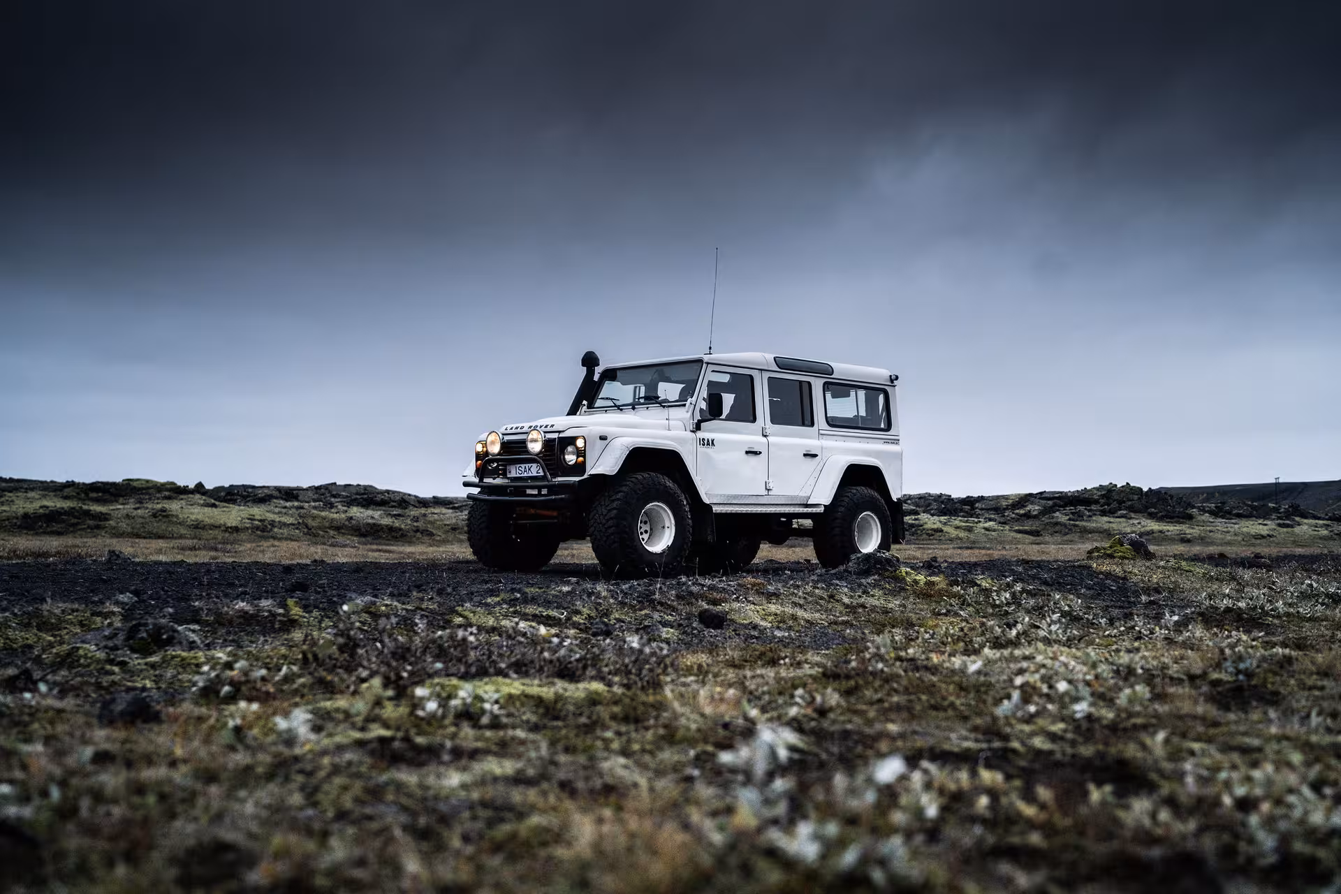 White Land Rover Defender with oversized tires on volcanic lava field landscape under dramatic sky