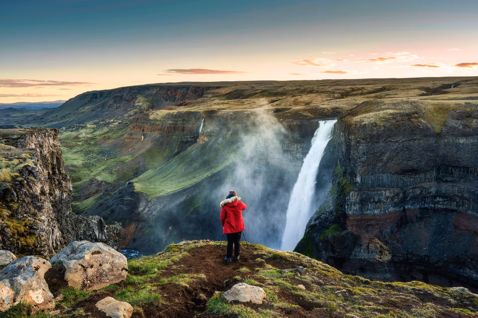 Photographer in red jacket on cliff edge capturing Háifoss waterfall in Iceland's highland canyon at sunset