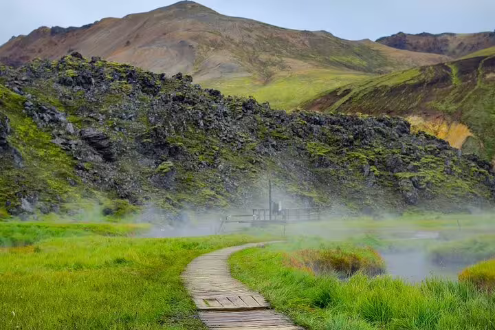 Wooden boardwalk path through steaming geothermal hot springs at Landmannalaugar with mossy lava fields and colorful rhyolite mountains