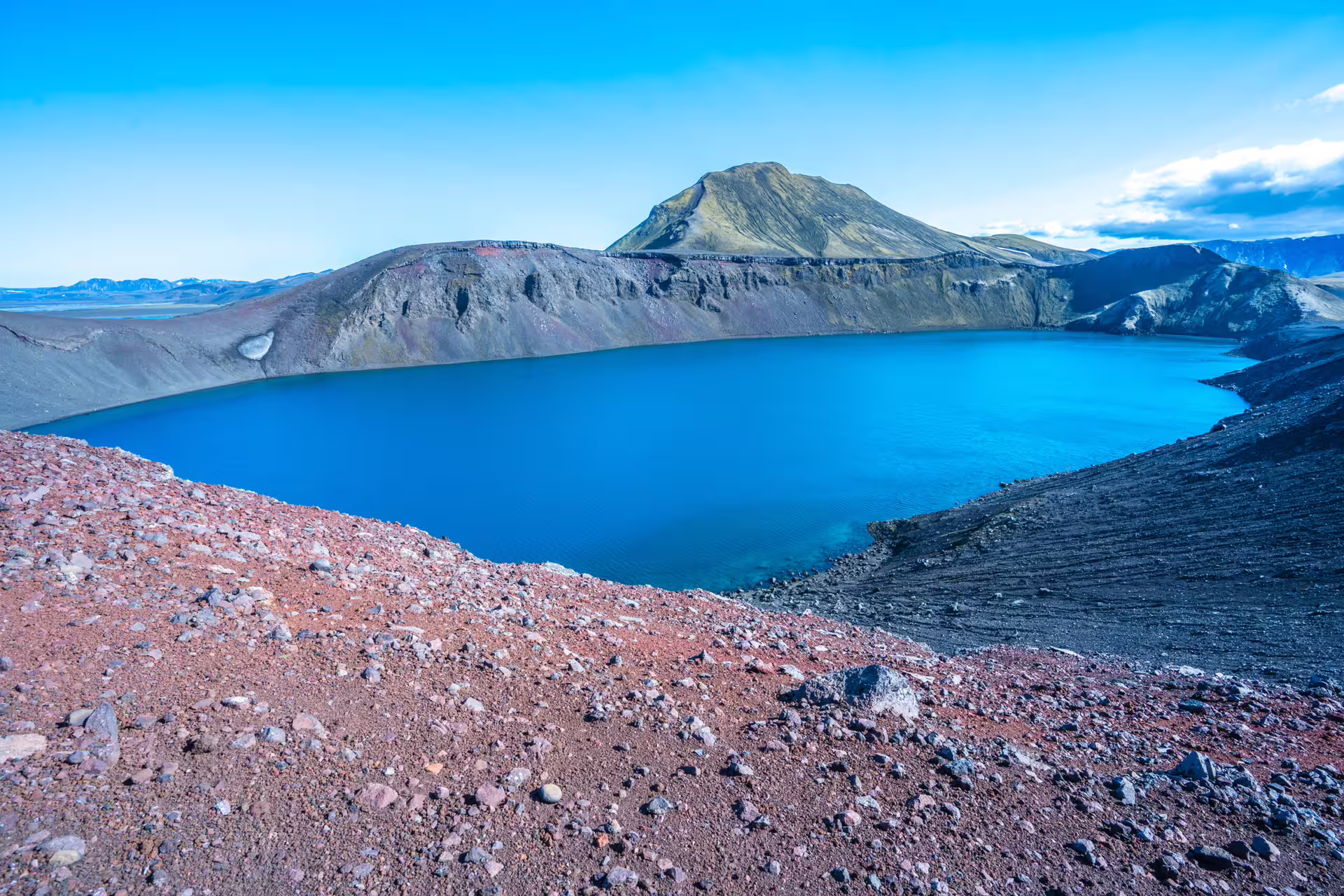 Vivid blue crater lake surrounded by red volcanic terrain and dark lava hills in Landmannalaugar, Iceland