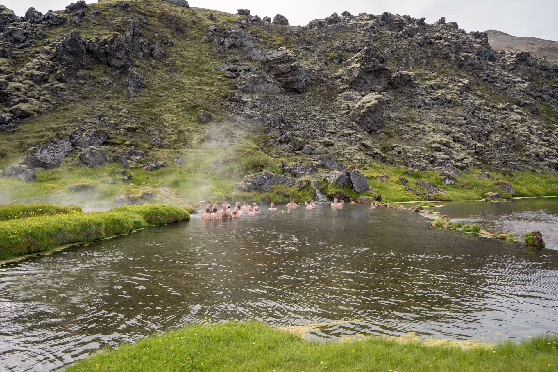 Group of people bathing in geothermal hot spring pool at Landmannalaugar with steam rising and volcanic landscape