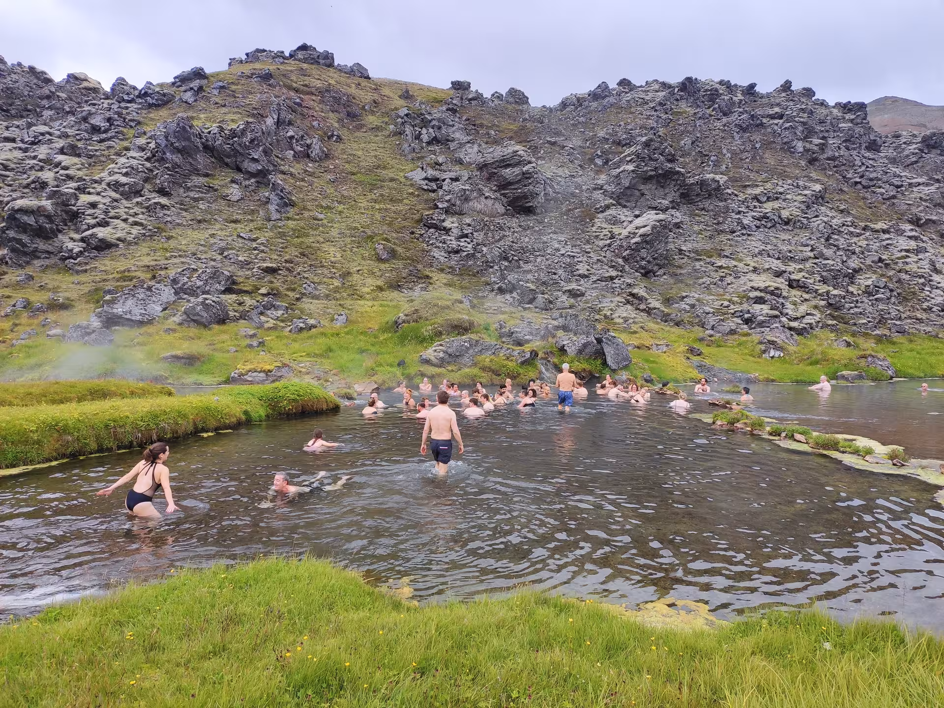 Tourists bathing in Landmannalaugar geothermal hot spring pool surrounded by volcanic rock and green hills