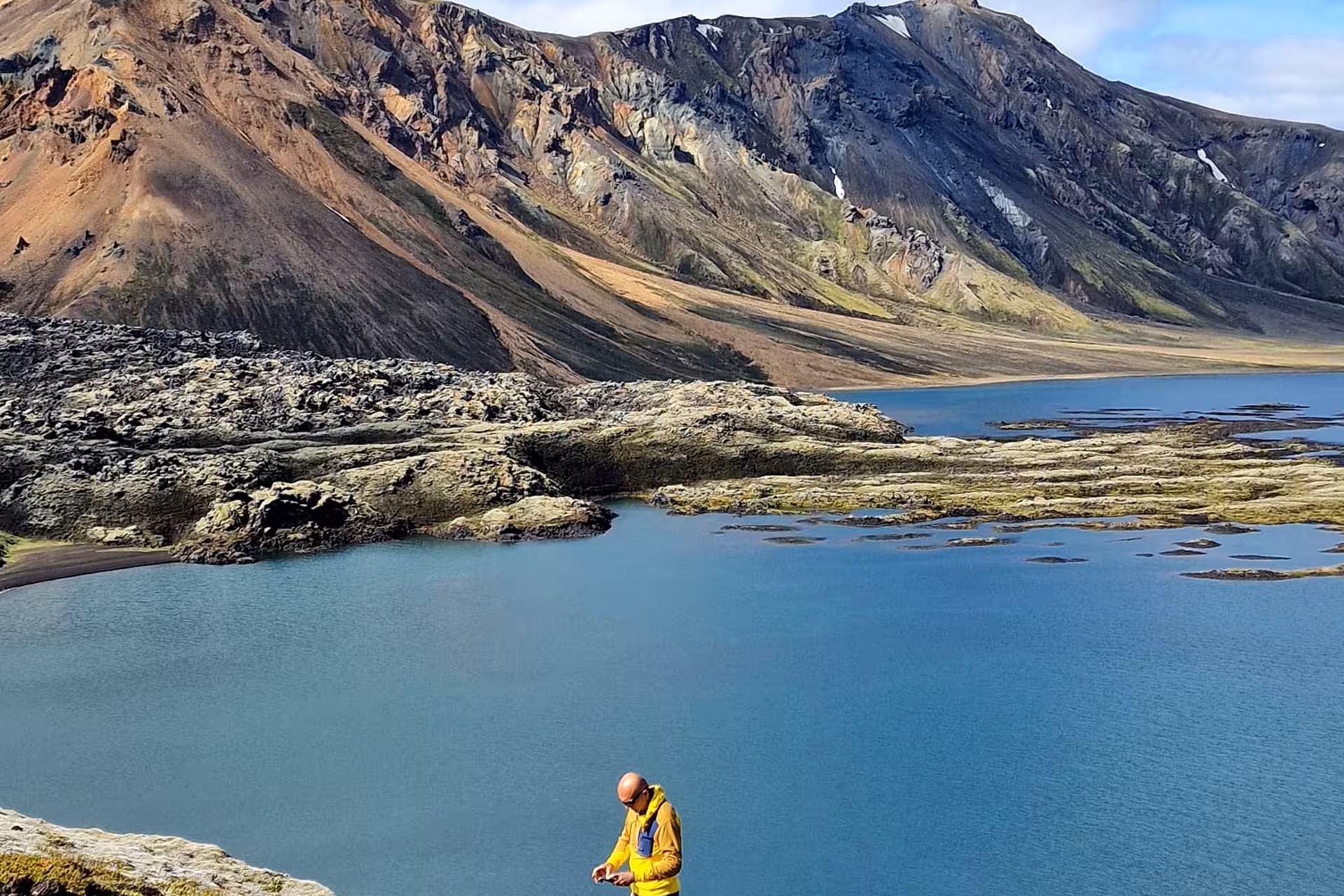 Hiker in yellow jacket at blue glacial lake surrounded by colorful rhyolite mountains in Landmannalaugar