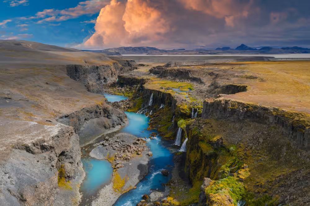 Turquoise river winding through Landmannalaugar canyon with waterfalls, mossy cliffs, and dramatic sunset clouds