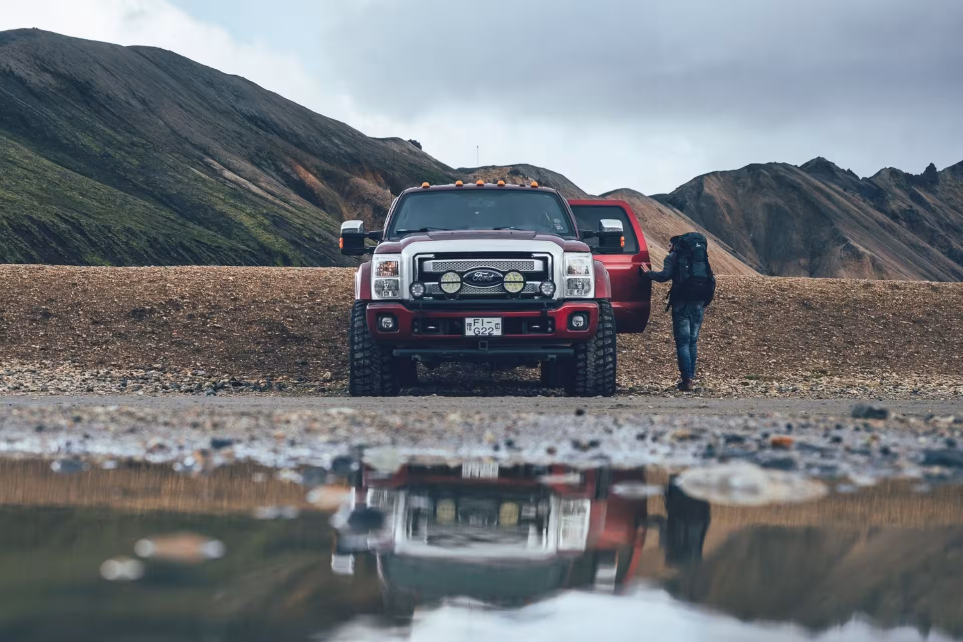 Red Ford pickup truck parked in Landmannalaugar highlands with hiker carrying backpack and colorful mountains behind