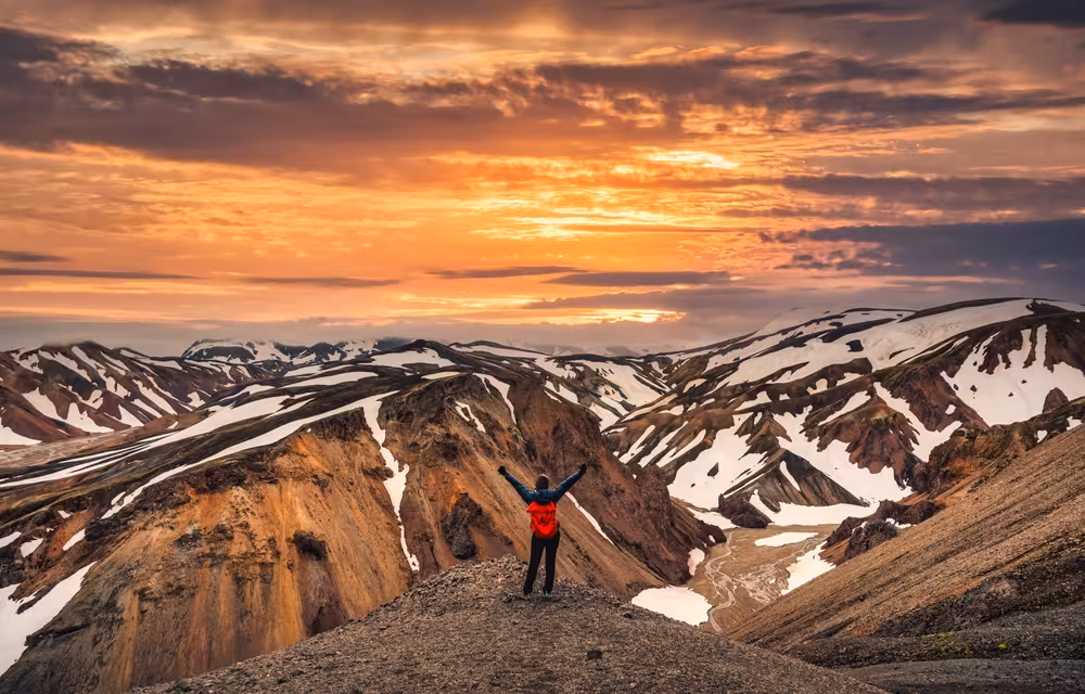 Hiker with arms raised overlooking snow-dusted colorful rhyolite mountains at sunset in Landmannalaugar highlands