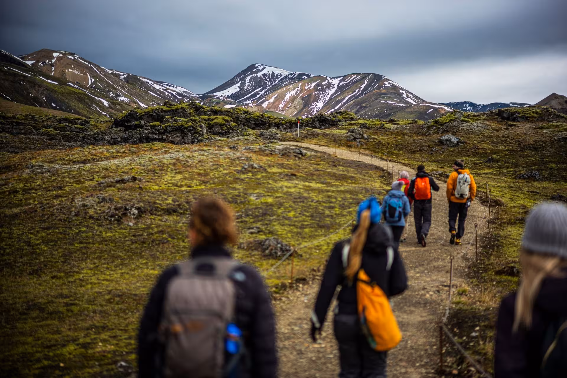 Hikers with backpacks walking a trail toward snow-capped Landmannalaugar highlands mountains under cloudy skies