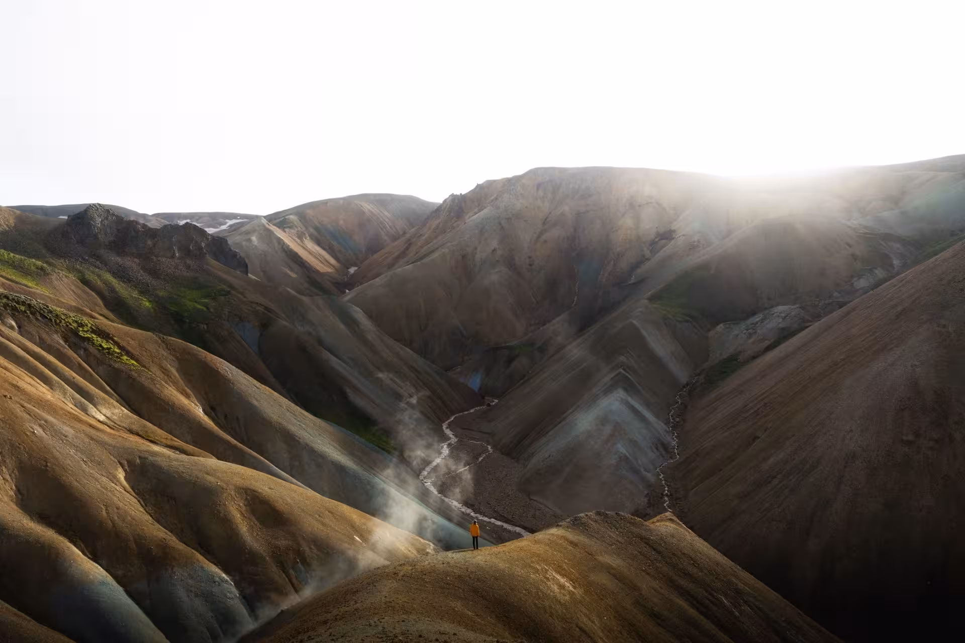 Lone hiker in yellow jacket standing on ridge overlooking colorful Landmannalaugar highland valley with geothermal steam