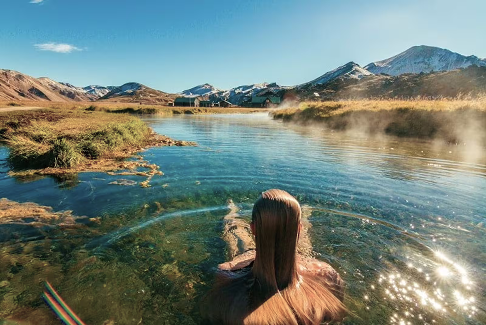 Woman bathing in steaming Landmannalaugar hot spring with snow-capped mountains and clear blue sky