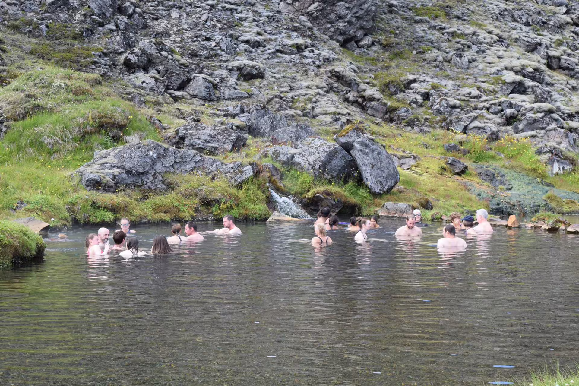 Visitors soaking in Landmannalaugar natural hot spring pool surrounded by volcanic rocks and green hills, Iceland