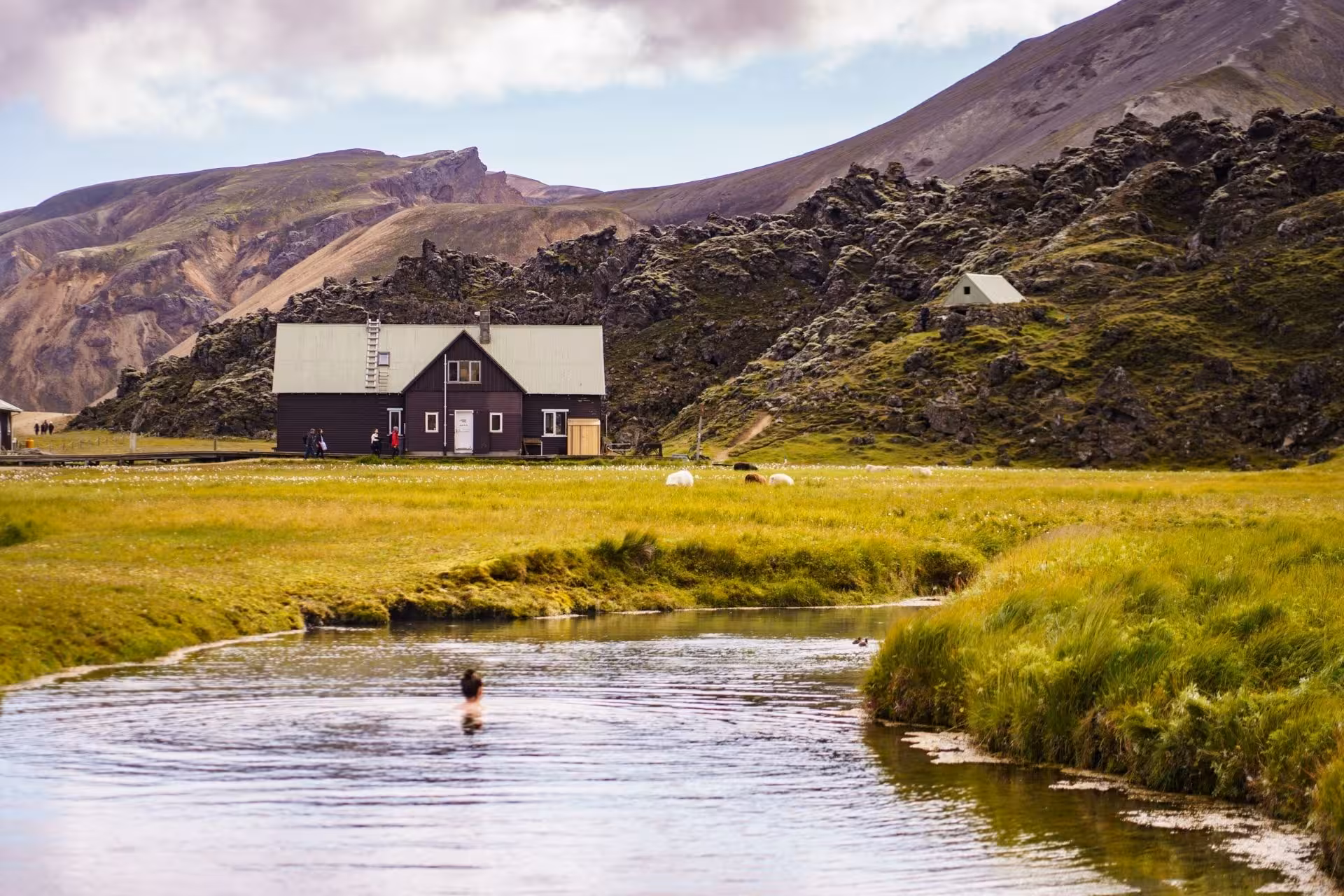 Person bathing in Landmannalaugar hot spring with dark wooden hut and volcanic highlands in background